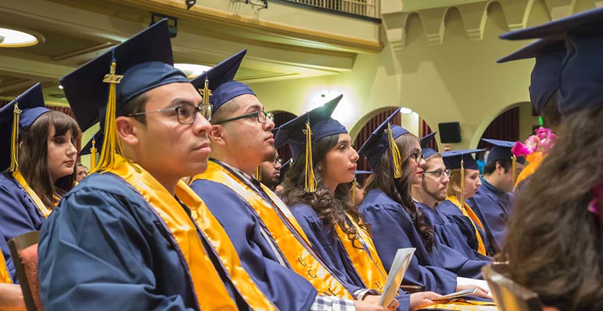 Graduates seated during commencement listen to a speaker on stage.