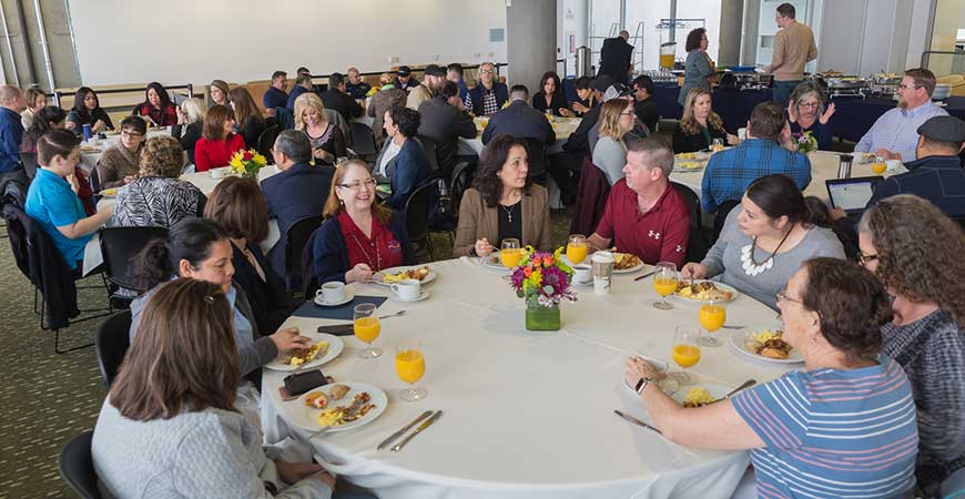 Several dozen UC Merced employees are seated at round tables eating breakfast in Room 355 of Kolligian Library.