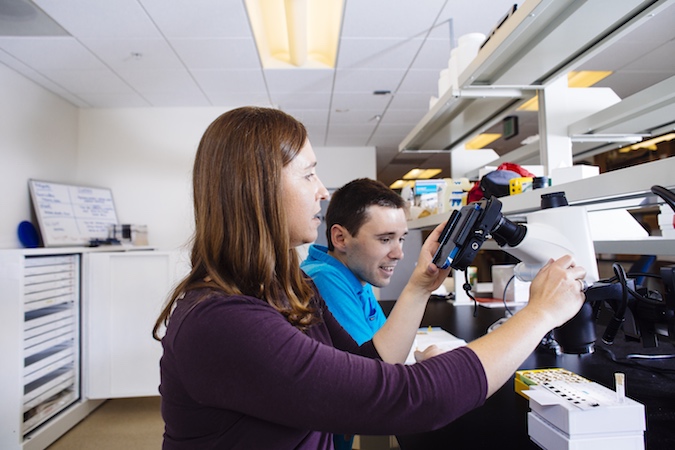 A woman and man sit in front of a microscope in a laboratory.