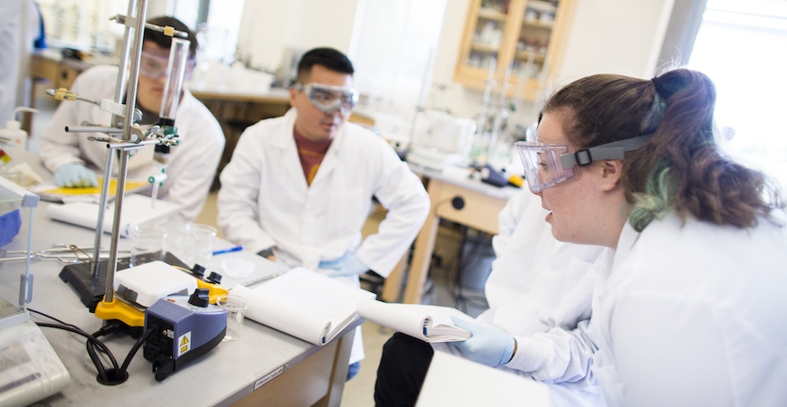 Students in white lab coats working at a lab bench with scientific equipment.