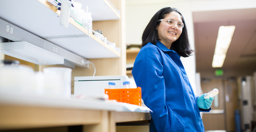 Professor Clarissa Nobile wearing a blue lab coat, teal-colored gloves, and safety goggles leans against a bench in her laboratory.