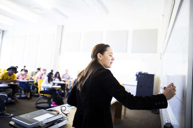 Professor Noemi Petra, pen in hand, writes on a white board in front a classroom full of students.