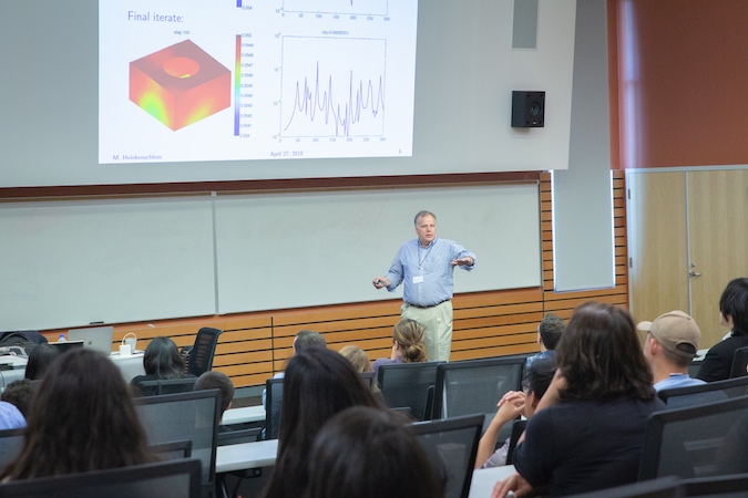 A man stands in front a projected image delivering a talk to a crowded lecture hall.